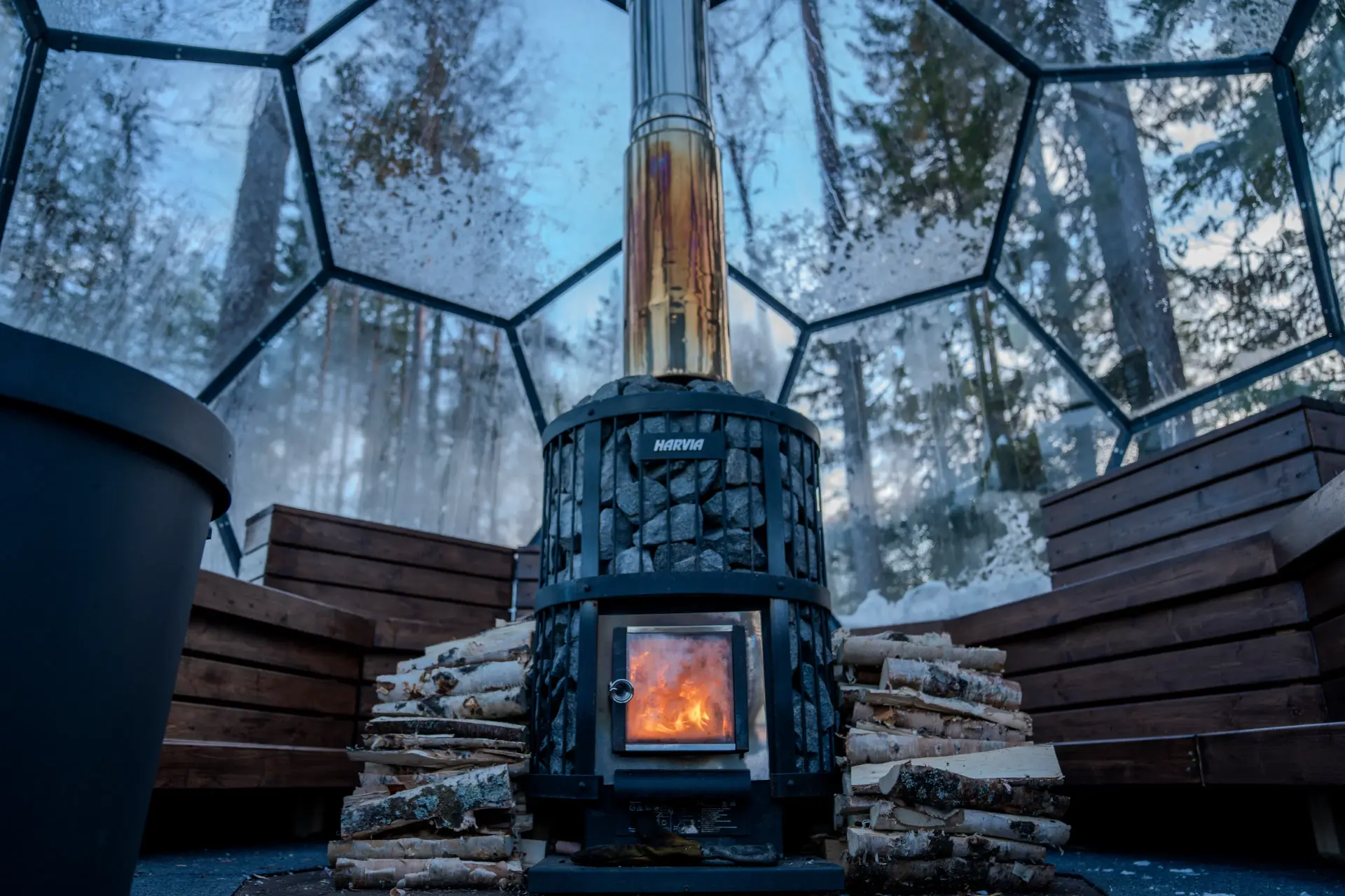 Wood-burning Harvia sauna stove with firewood inside the glass igloo surrounded by snowy forest