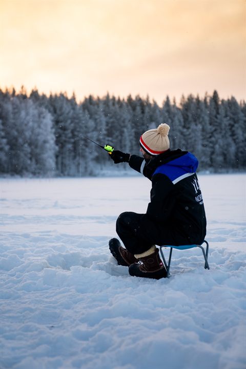 Guest ice fishing on a small stool on the frozen lake, snowy forest behind