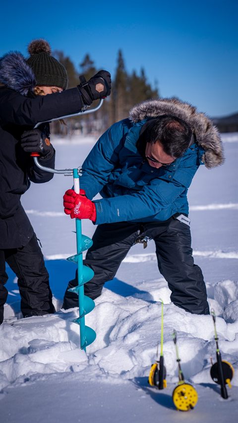 Two guests drilling a hole in the lake ice with an ice auger, fishing rods ready in the snow