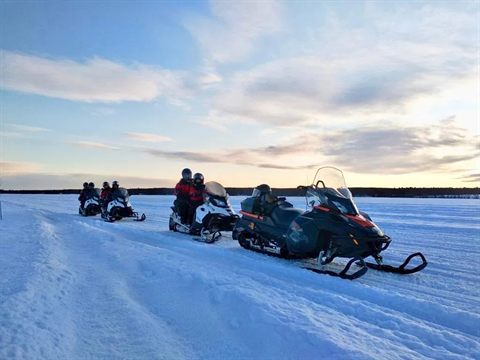 A row of snowmobiles with riders on a frozen lake at dusk