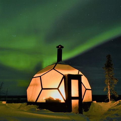 Glasiglu-Sauna leuchtet warm unter einer grünen Aurora in einer verschneiten Nacht