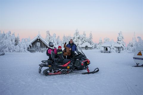 A family with two children by a snowmobile in front of snow-covered wilderness huts