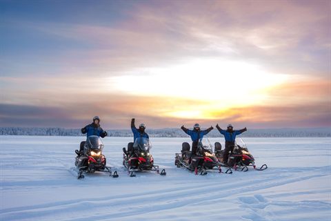 Four snowmobile drivers cheering on a frozen lake at sunset
